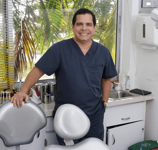 Doctor Jairo Montoya MD - Dentist Cartagena Dr. Jairo Montoya smiling next to his dentist's chair with palm trees outside the window.